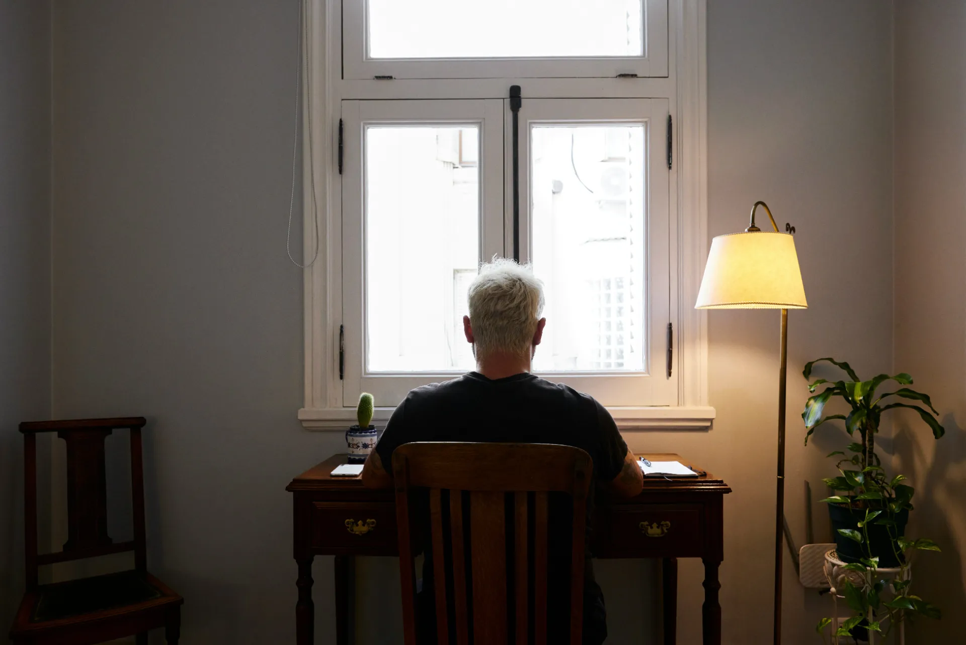 Older adult sitting alone at a desk by a window in a dim room, showing depression and isolation